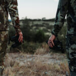 Two men in camouflage holding guns while standing on a hill.